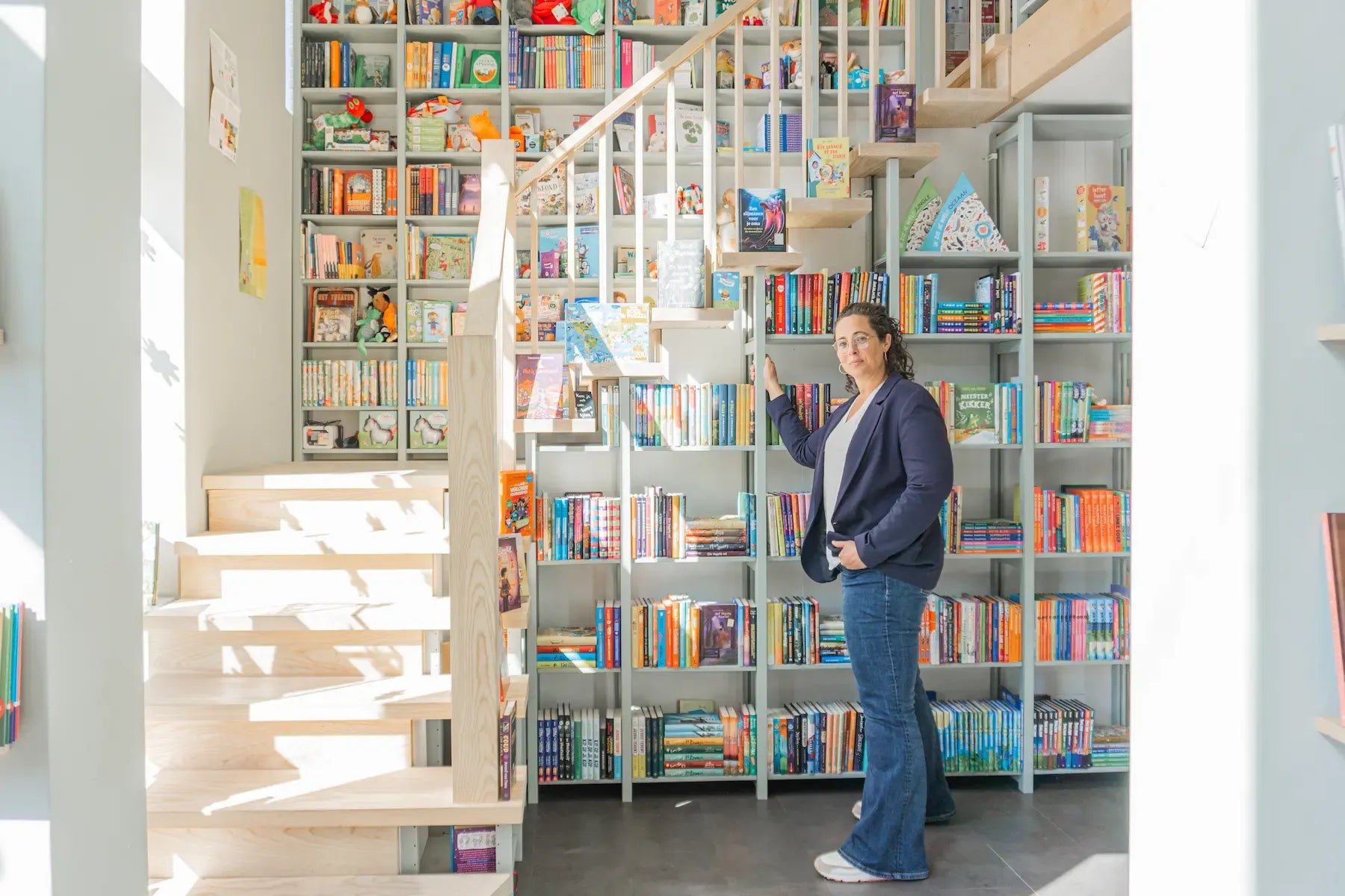 Eine Frau steht neben einem hohen, weißen Bücherregal voller bunter Bücher neben einer hellen Holztreppe in einem sonnigen Zimmer.