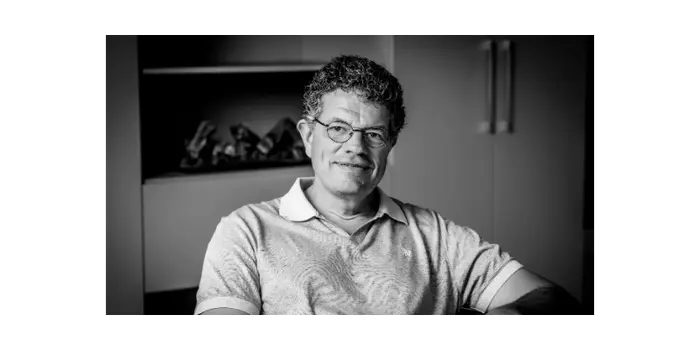 Zwartwit portret van een man met bril en gekruld haar zittend in een kamer met kasten en decoraties op de achtergrond.