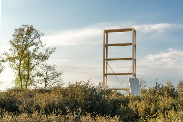 Hölzerner Aussichtsturm auf einem Feld mit Büschen unter klarem Himmel mit vereinzelten Wolken.