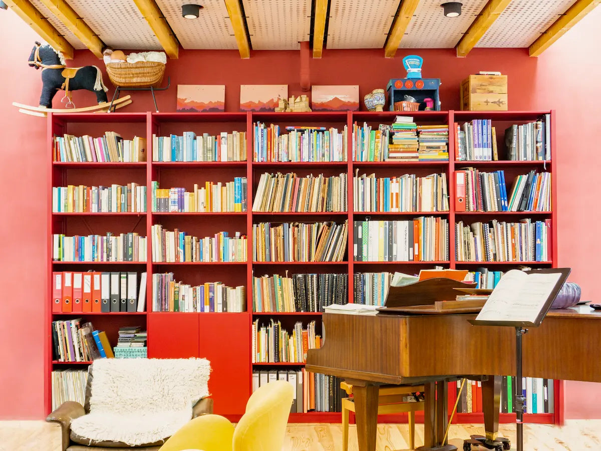 Grote rode boekenkast gevuld met diverse boeken, houten piano en muziekstandaard in warme woonruimte met houten plafondbalken
