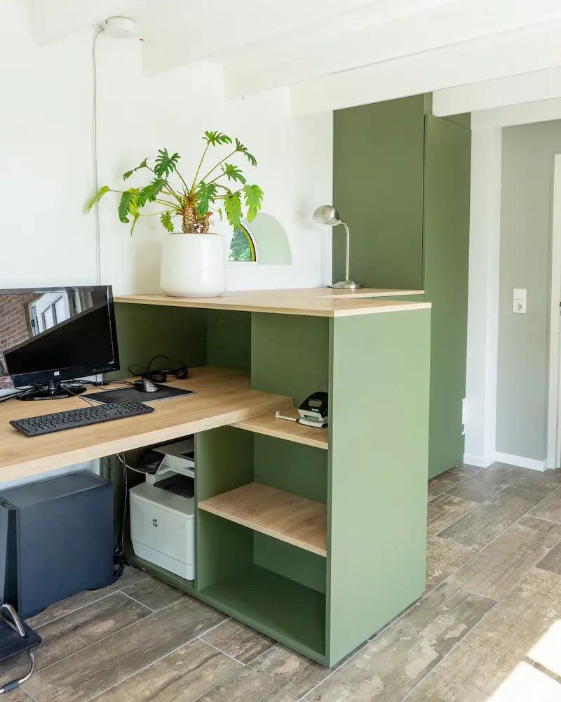 Hoekwerkplek met licht houten blad, groene kast met open vakken en een witte pot met grote kamerplant erop.