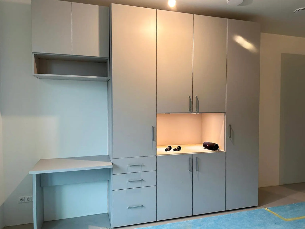 Wandvullende lichtgrijze kast met verschillende deuren en lades, open vak met verlichting en werkblad aan de zijkant.