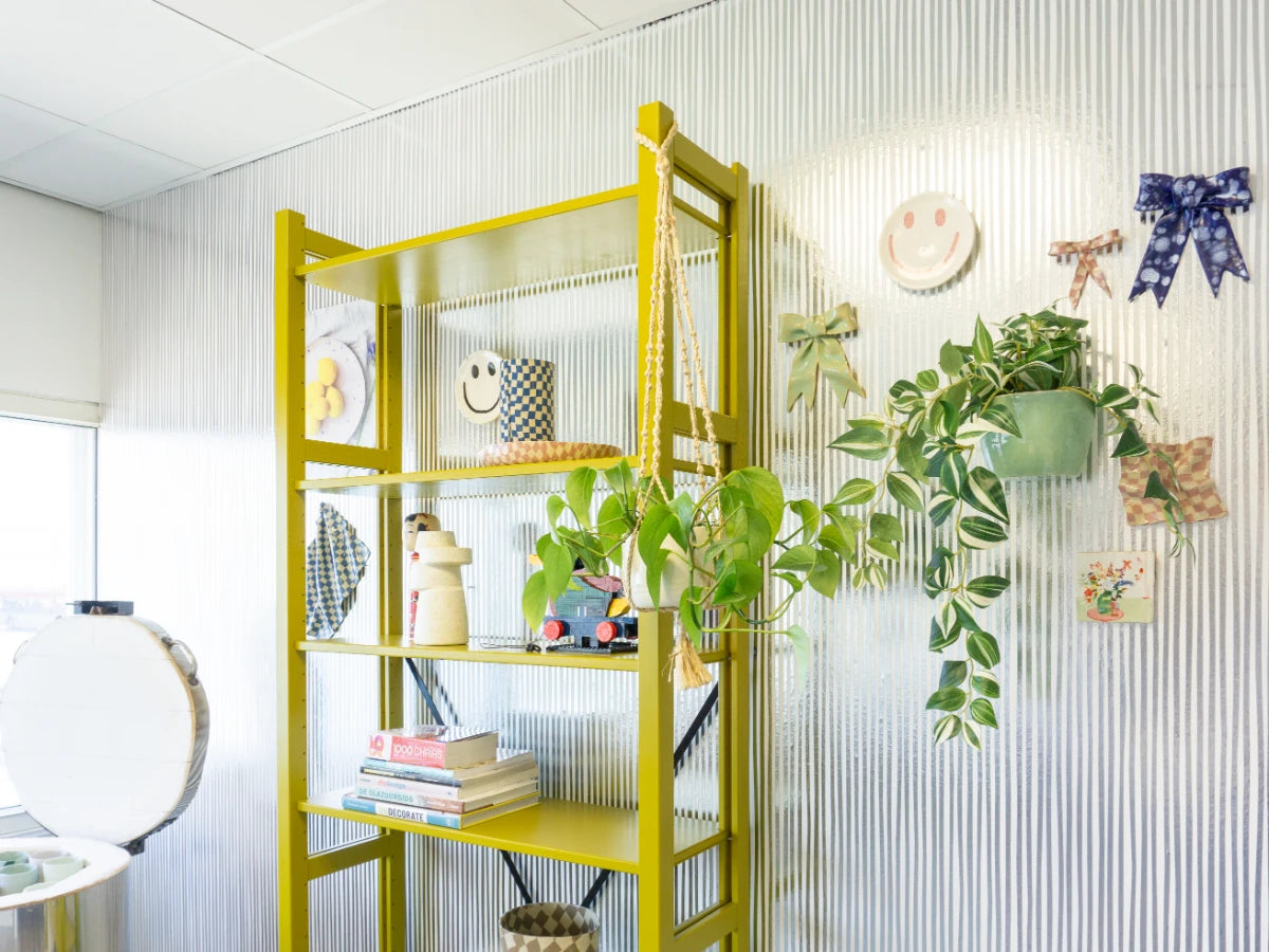 Lichte hoek met gele open kast vol planten, boeken en decoraties naast wand met kleurrijke strikken en smiley afbeeldingen