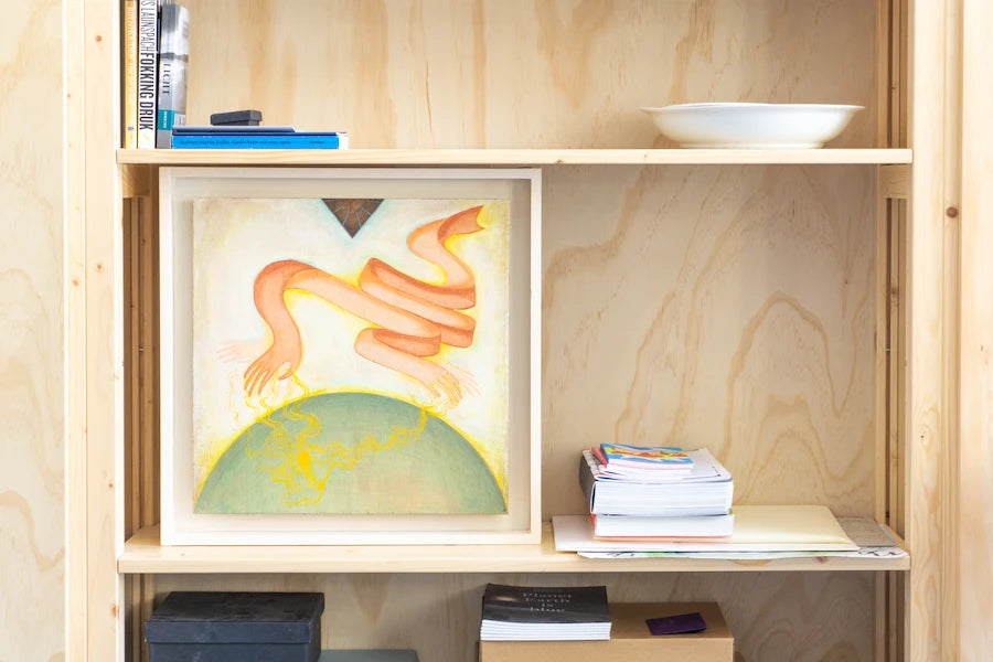 Open houten kast met kleurrijk abstract schilderij, stapels papier en enkele boeken tegen lichte houten achtergrond