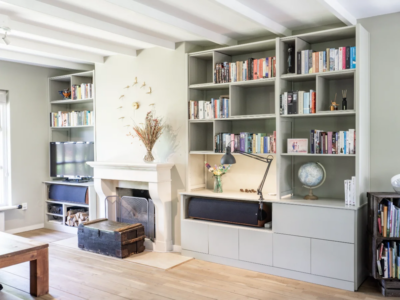 Helles Wohnzimmer mit grauen Wandschränken voller Bücher, Kamin, Holzboden und gemütlichem Arbeitsbereich mit Schreibtischlampe