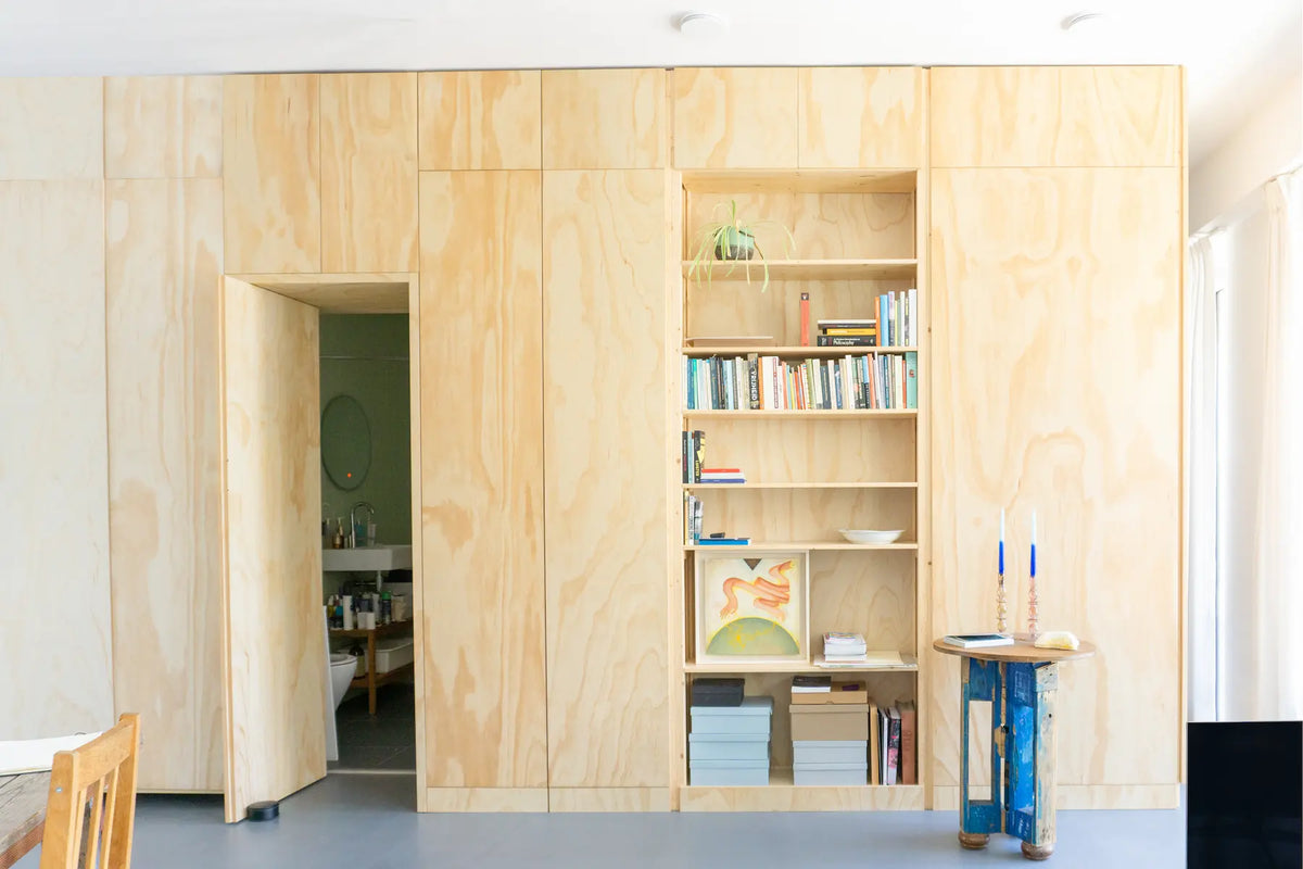 Lichte houten wand met open vakken gevuld met boeken en decoraties naast draaibare deur en blauwe tafel met kaarsen