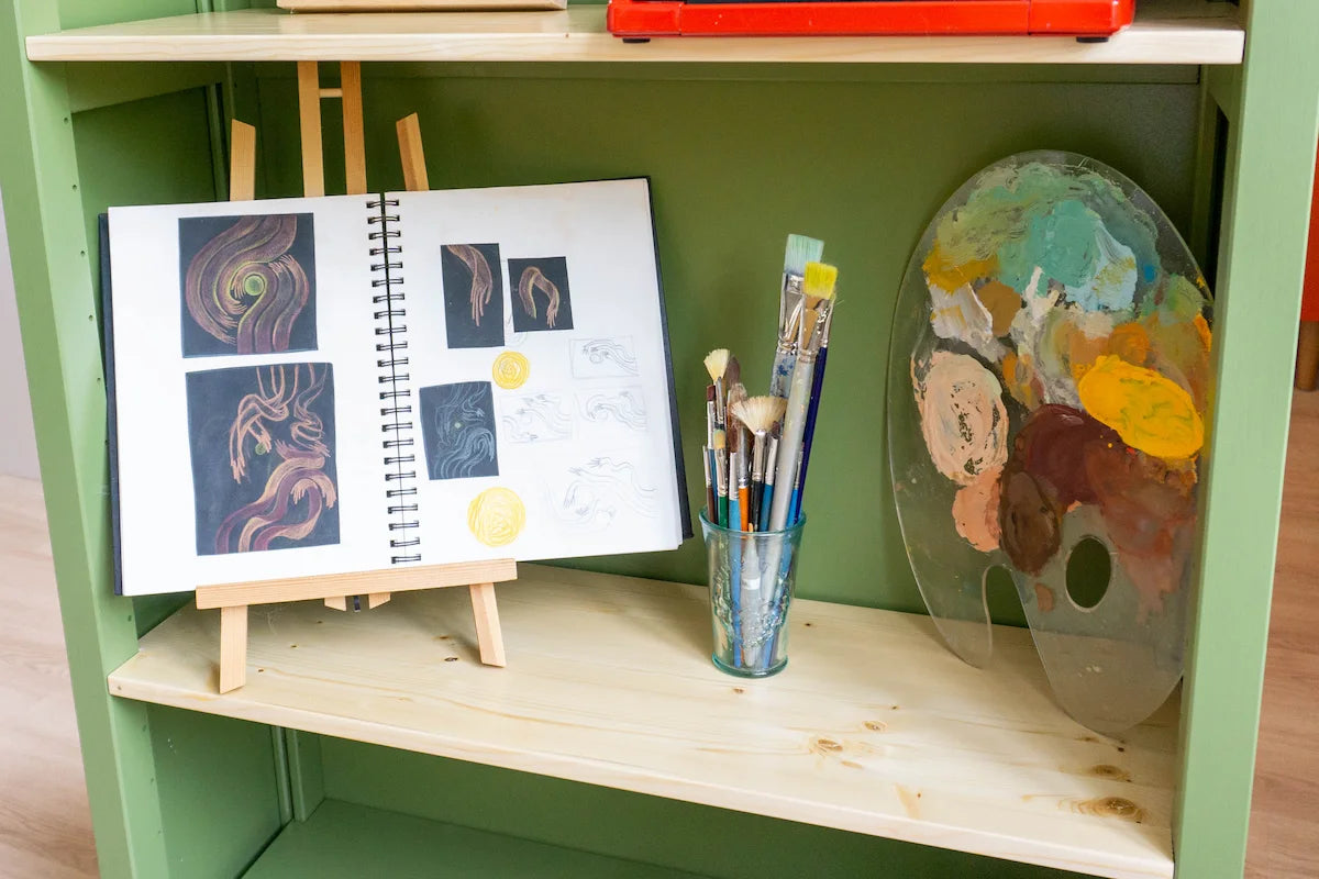 Open schetsboek met tekeningen op ezel, glazen pot met kwasten en beschilderd palet in groen houten kastje