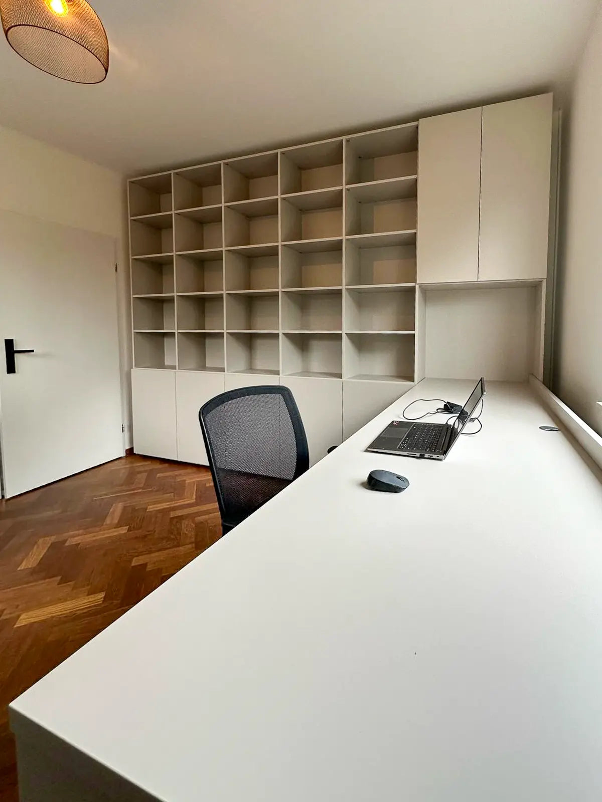 Witte werkplek met lang bureau, zwarte bureaustoel en grote open kast met vakken en bovenkastjes tegen muur.